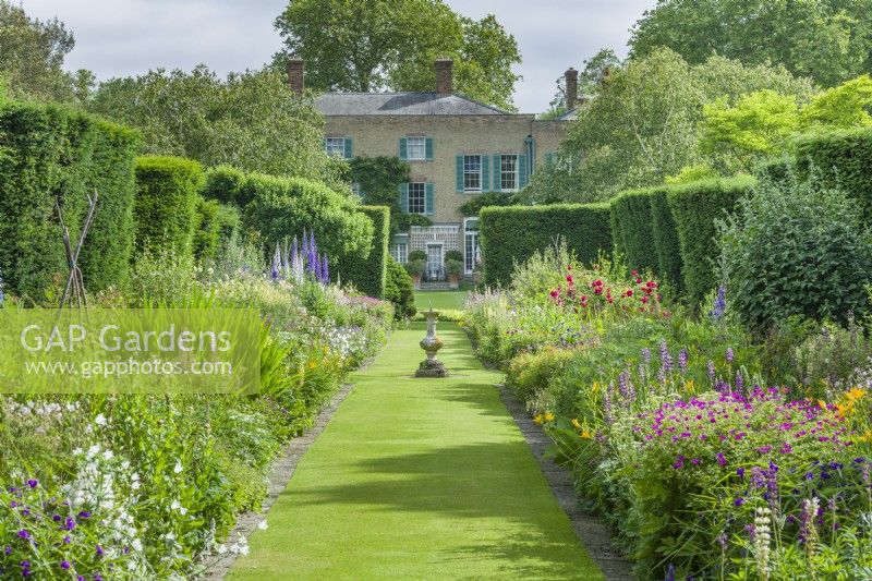 View of double herbaceous borders and Abbots Ripton Hall, June. Grass paths and sundial at intersection.