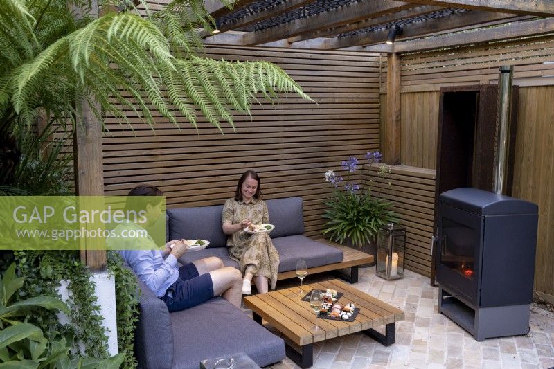 Man and woman eating food on garden seat in contemporary wood pergola with log burner