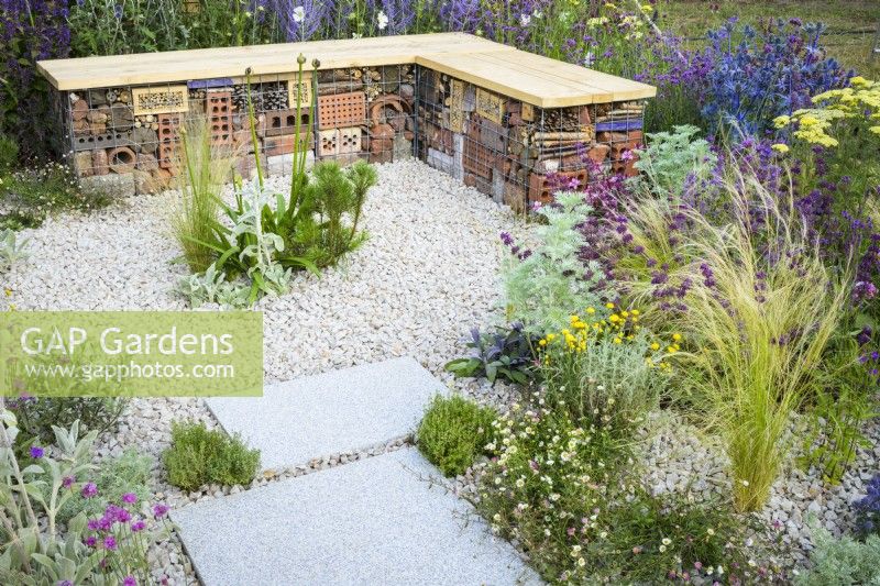Bug or insect hotel inside gabion bench with wooden  top in low maintenance gravel garden with drought tolerant plants to attract pollinators, such as Salvia, Achillea, Stachys byzantina, Eryngium, Santolina and Stipa tenuissima- Turfed Out, RHS Hampton Court Palace Garden Festival 202