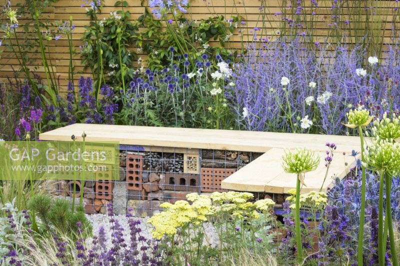 Bug hotel gabion bench with wooden  top in low maintenance gravel garden with drought tolerant plants to attract pollinators, such as Salvia, Achillea 'Credo', Agapanthus, Stachys byzantina and Stipa tenuissima -Turfed Out, RHS Hampton Court Palace Garden Festival 2022

