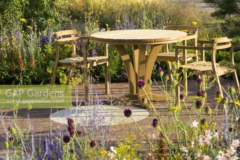 Sitting area with wooden table and chairs and planting of  Allium sphaerocephalon, Achillea 'Coronation Gold'  and Perovskia in Joy club garden - RHS Hampton Court Palace Garden Festival 2022