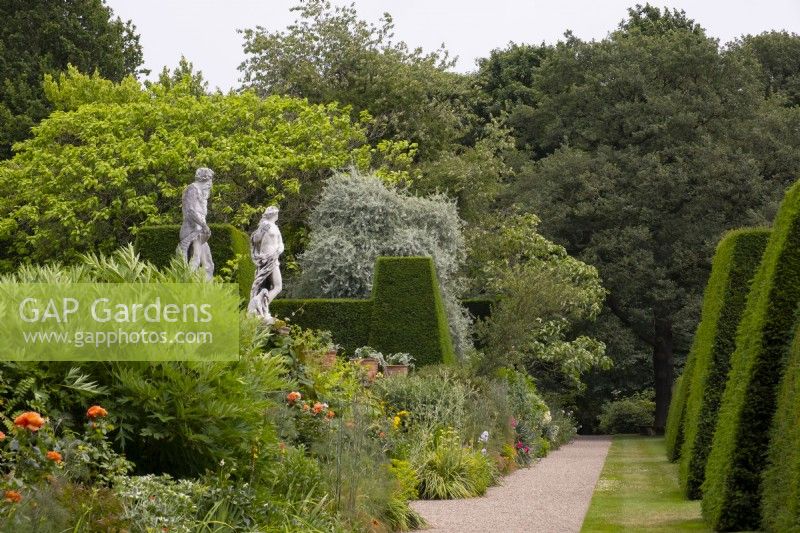 Stone statues, herbaceous borders and Taxus baccata topiary along a gravel path at Renishaw Hall and Gardens.