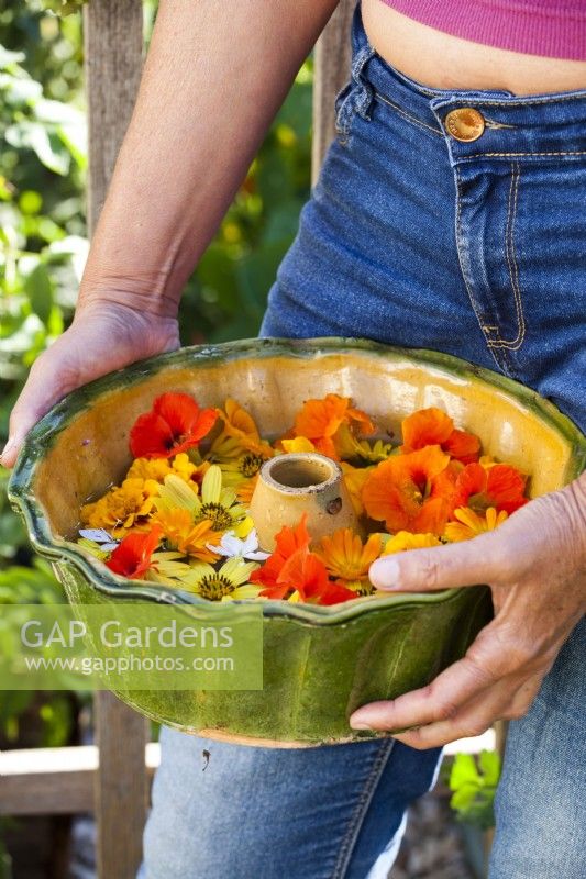 Woman holding cake pan with cut flowers including nasturtium, pot marigold, French marigold, Gaura and Helianthus ' Lemon Queen'.