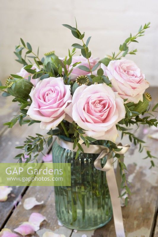 Pink roses arranged with eucalyptus and papaver seedheads in a blue glass jar tied with a ribbon against rustic background
