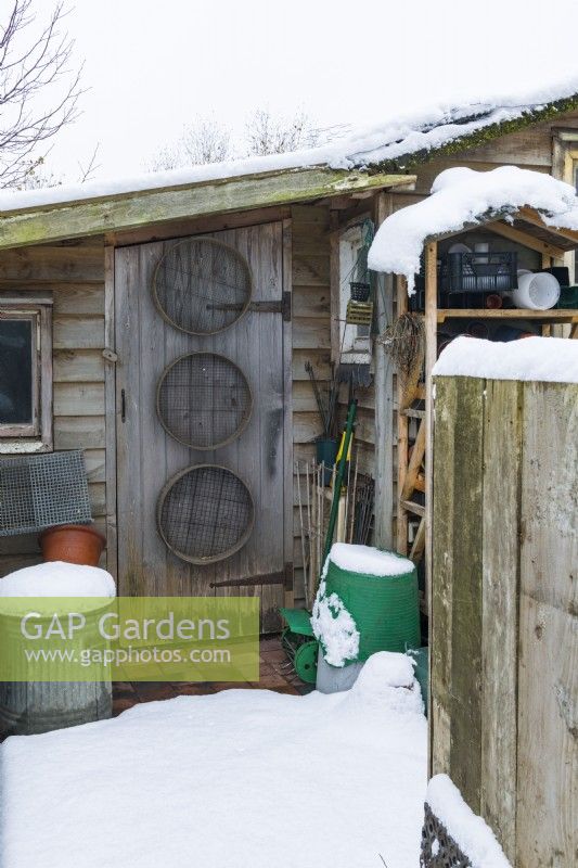 Small leanto garden shed and well-organised covered storage area with garden sieves, tools and containers. Snow. December.