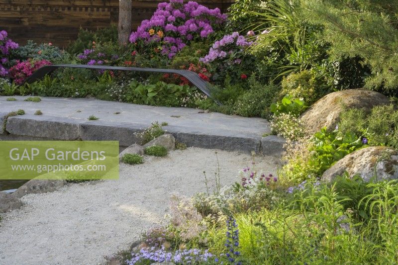 Boulders among planting with bespoke iron bench overlooking a pond with colourful Rhododendrons behind - A Swiss Sanctuary, RHS Chelsea Flower Show 2022