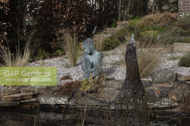 A cormorant sculpture made of recycled wire and mesh in front of image beside a pond, with Marissa, a copper bronze sculpture by Jenny Wynne Jones, sat amongst gravel and grasses in background. February. 
