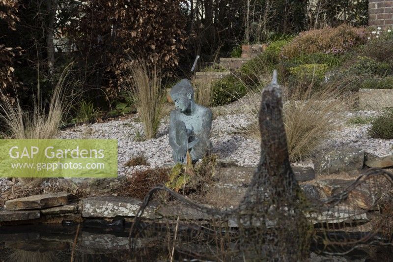 A cormorant sculpture made of recycled wire and mesh in front of image beside a pond, with Marissa, a copper bronze sculpture by Jenny Wynne Jones, sat amongst gravel and grasses in background. February. 