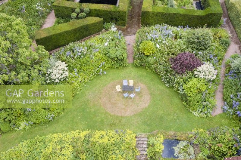 View over wide border of Alchemilla mollis and mixed border surrounding lawn with metal table and chairs painted pastel colours and formal hedges of Yew in Background. Image taken from drone. July.