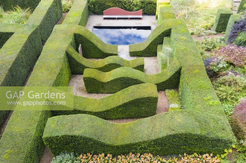 View over clipped mature hedges of Yew containing a formal rectangular reflecting pool and seat and a line of hedges separated by gravel paths. July. Image taken from drone. 