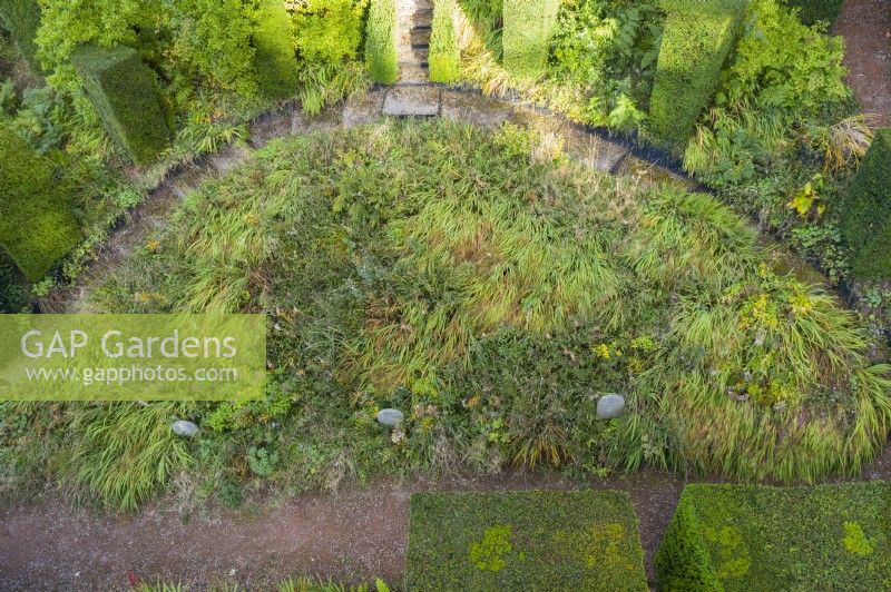 View over semi circular shaped border contained by gravel paths and surrounded by tall columns of clipped Yew. Main border is of herbaceous plants grown 'wild' without active cultivation. Image taken from drone. July.