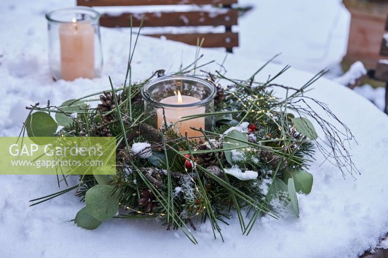 A pillar candle in storm glass surrounded by wreath decorated with pine needles and branches and Eucalyptus foliage on a snowed table
