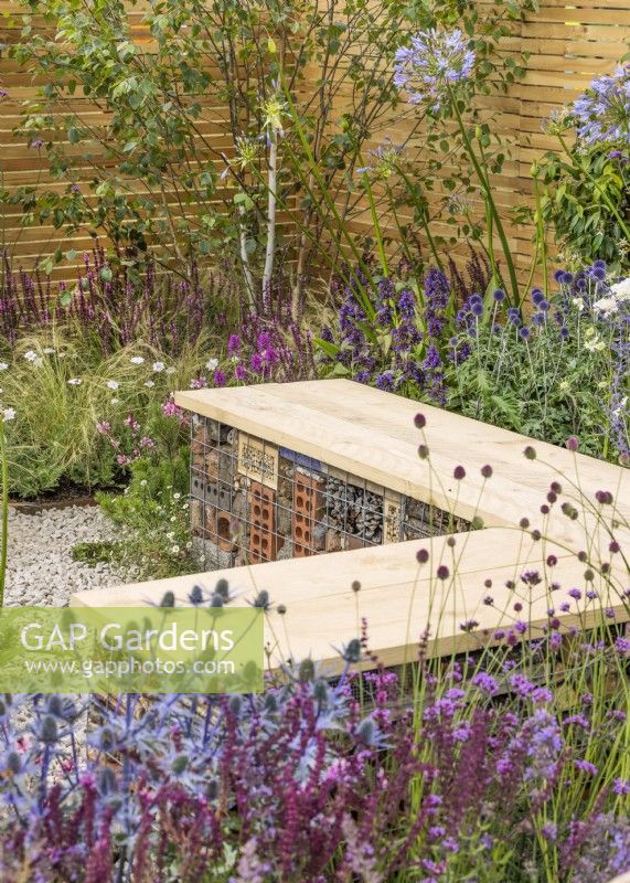 L-shaped wooden bench incorporating 'bug hotel' underneath, surrounded by drought resistant plants in gravel, summer July