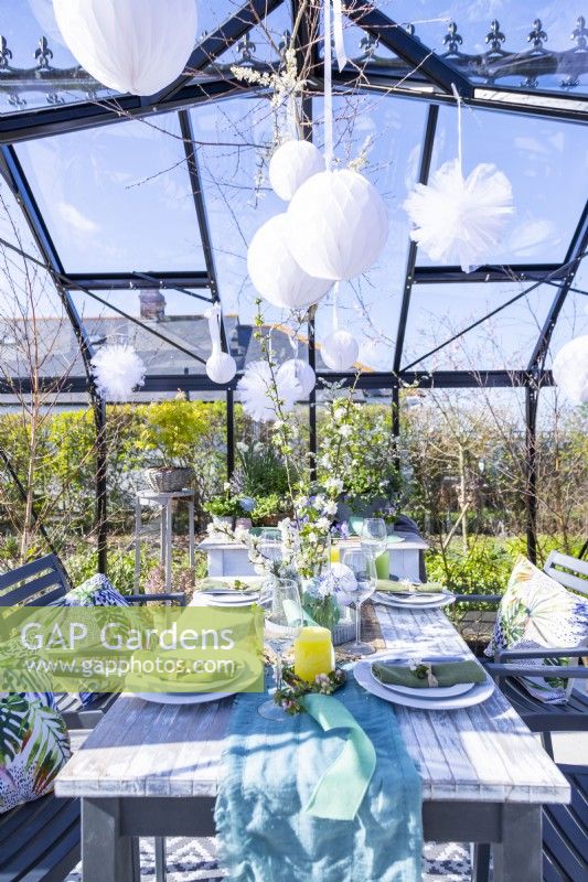 Table set out in greenhouse with plates, cutlery, glasses, candles and a flower display in the middle and hanging paper pom poms