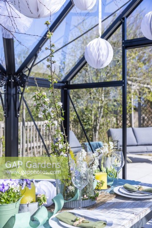 Table set out in greenhouse with plates, cutlery, glasses, candles and a flower display in the middle and hanging paper pom poms