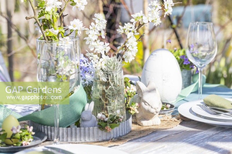 Table set out in greenhouse with plates, cutlery, glasses, candles and a flower display in the middle with easter decorations