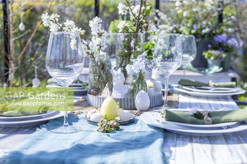 Table set out in greenhouse with plates, cutlery, glasses, candles and a flower display in the middle with easter decorations