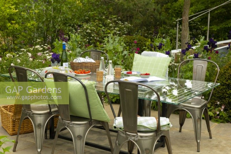 A glass dining table and metal chairs surrounded by colourful borders in Cavernona on My Mind, a sanctuary garden designed by Taina Suonio and Anne Hamiltion at the RHS Chelsea Flower Show 2023.
