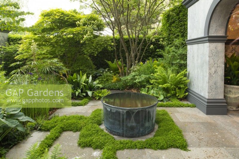 A circular metal water feature surrounded by ferns, trees and Yew hedge in the Myeloma UK - A Life Worth Living Garden designed by Chris Beardshaw at the RHS Chelsea Flower Show 2023