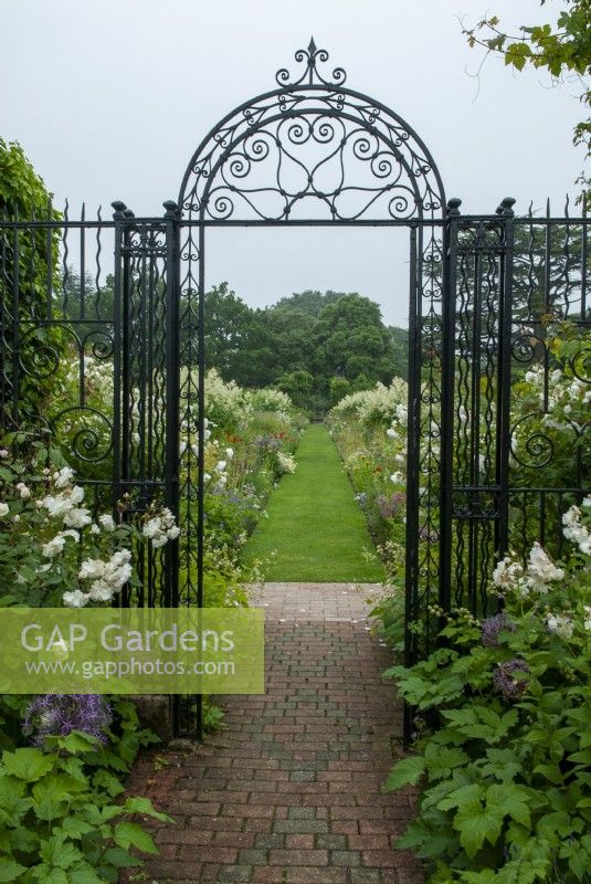 Decorative wrought iron entrance gate to classical 16th century garden, with view to grass path and deep herbaceous borders on either side  - The Walled Garden, Helmingham Hall, Suffolk