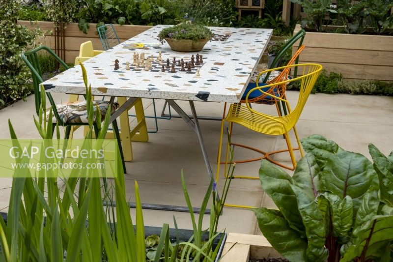 A community table that incorporates two chess boards and is place for people to come together to eat or play games. Plants in the foreground, red chard and Iris pseudacorus with a terra cotta bowl of Thymus vulgaris. The London Square Community Garden, Gold winner - Chelsea 2023 Designed by: James Smith