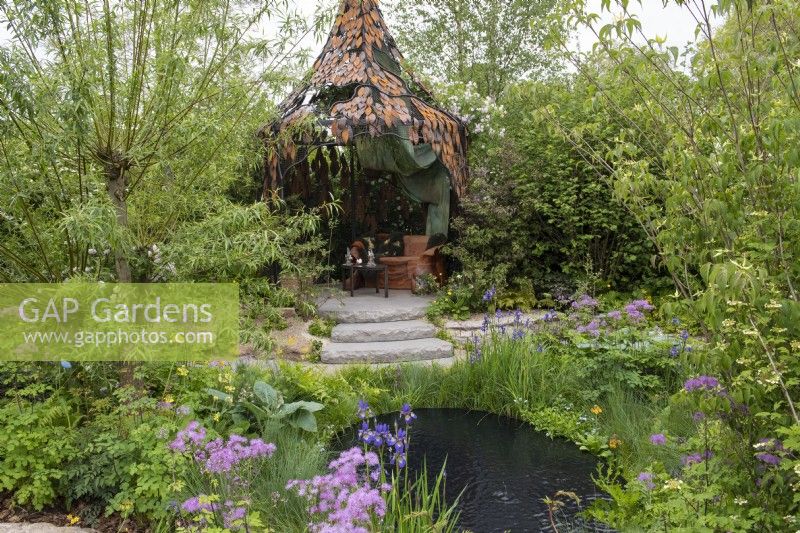A forged iron pavilion crafted by Cox London overlooks a pool edged in Salix alba, Iris setosa var. arctica, thalictrum,  candelabra primulas, hostas and fern