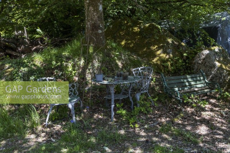 An old set of wrought iron metal table and chairs in dappled shade under a tree beside a bank with a few old plant pots on the table. Dartmoor. May. Spring. 