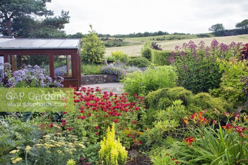 Monarda 'Gardenview Scarlet', hemerocallis, eupatorium etc by conservatory; rolling countryside beyond