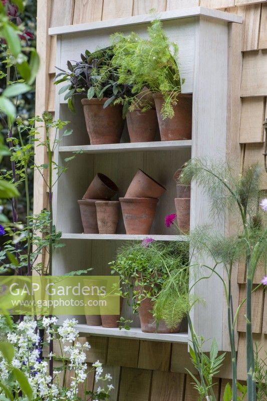A small shelving unit with terracotta pots and herbs is fixed onto a wall clad in cedar roof shingles. 