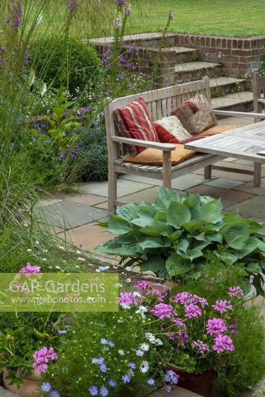 Sunken patio with table, seat and cushions and raised borders of perennial plants. Steps lead up to lawn at higher level - Open Gardens Day, Tuddenham, Suffolk



