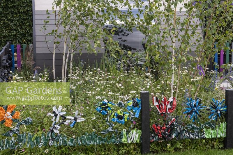 A playful way of upcycling waste plastic bags by weaving them into the chickenwire fence. Behind, a birch grove and meadow of wildflowers and grasses.