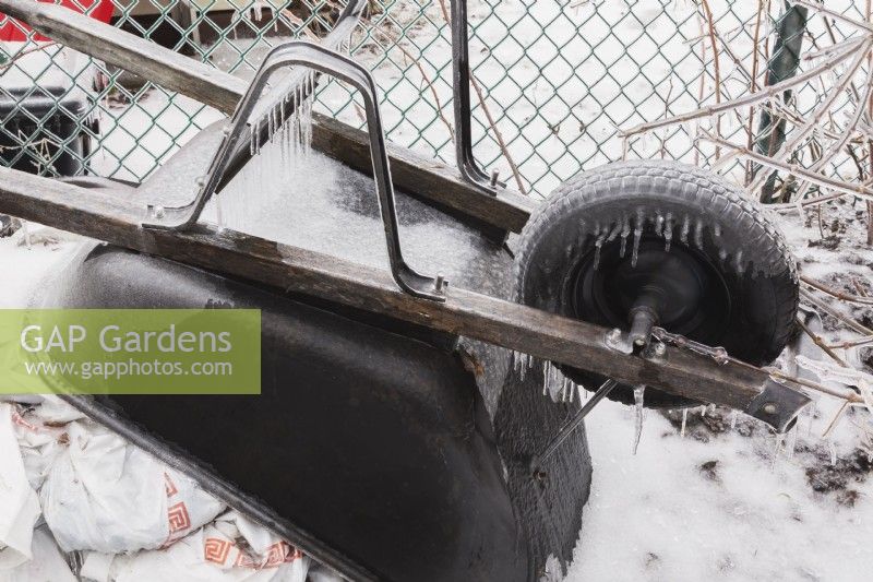 Stored black metal wheelbarrow covered in ice in backyard after freezing rainstorm in early spring.