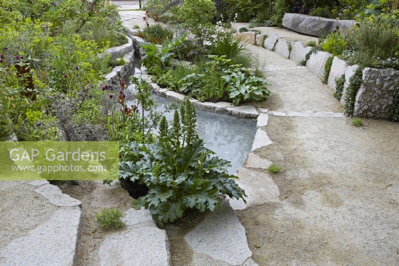 The Samaritans Listening Garden. Designer: Darren Hawkes. Chelsea Flower Show 2023. A garden of salvaged materials showing steps, path and raised bed edges near water. Summer. May.