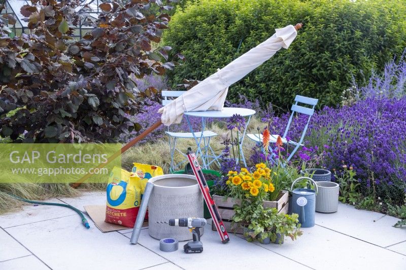 Parasol, Postcrete, scaffolding pole segment, sprit level, drill, duct tape, hosepipe, watering can and mixed plants laid out on ground