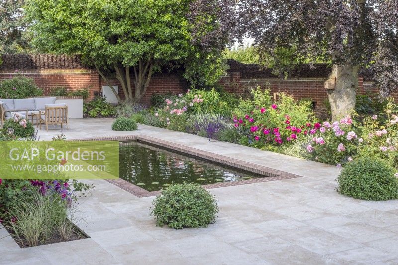 Brick edged rectangular pool on terrace of sandstone paving with garden furniture.  Borders include David Austin roses; Salvia nemorosa 'Caradonna', lavender, Ballota and Pittosporum topiary balls