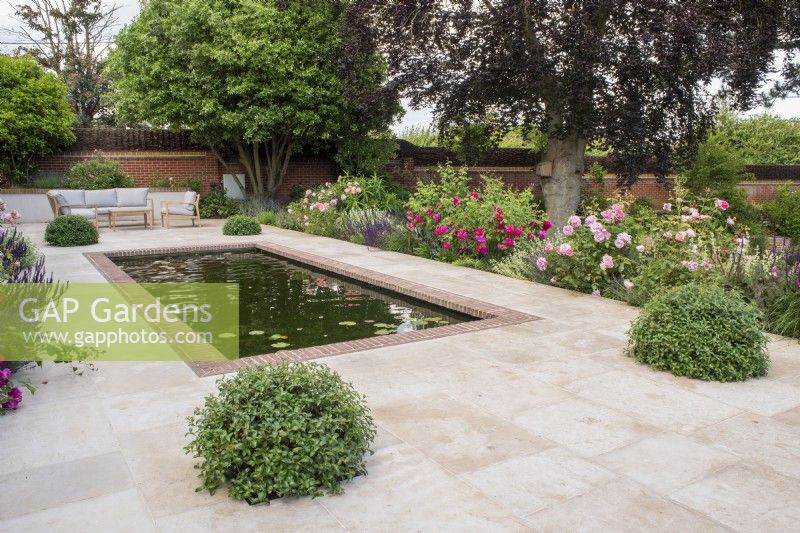 Brick edged rectangular pool on terrace of sandstone paving with garden furniture.  Border plants include David Austin roses; Salvia nemorosa 'Caradonna', Lavender, Ballota and Pittosporum topiary balls