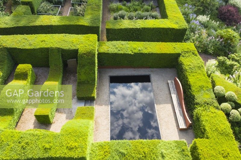 Clipped formal hedges of Taxus baccata - Yew, forming garden rooms, one containing a pool with dramatic reflections of the cloudy sky and a large bench. Image taken with drone. June. Summer. 