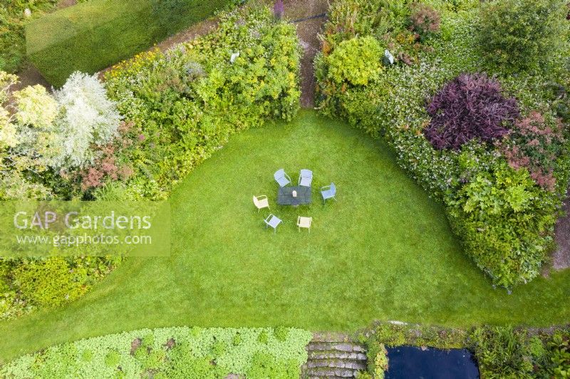 View over crescent shaped border of mixed shrubs and late perennials. Lawn with painted metal table and chairs. September. Image taken with drone. 