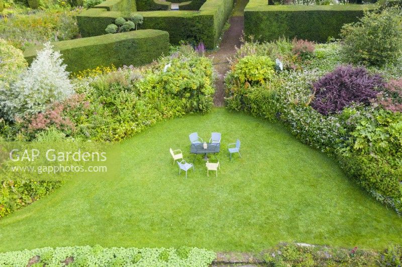 View over crescent shaped border of mixed shrubs and late perennials. Lawn with painted metal table and chairs. September. Image taken with drone. 