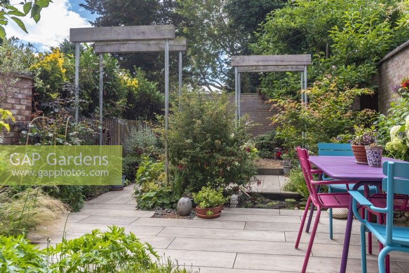 Small paved urban garden featuring recycled single pergolas with scaffold poles and half oak sleepers; pool and colourful table and chairs.  