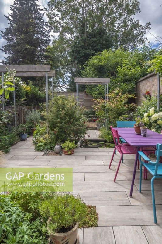 Small paved urban garden featuring industrial style single pergolas with scaffold poles and half oak sleepers; pool and colourful table and chairs.  Plants inc: Acers, Fuchsias and perennials