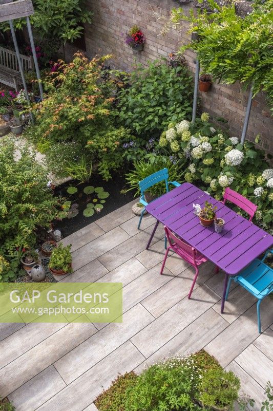 Birds eye view down on small enclosed paved urban garden featuring patio,containers, rectangular pool with waterlilies, colourful table and chairs. Planting inc: Hydrangea 'Annabelle', Acers, Wisteria and Fuchsias