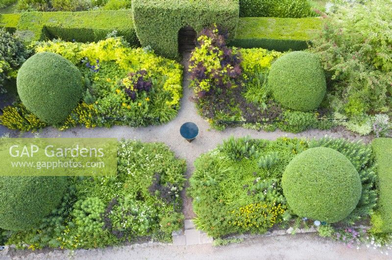 View over small formal garden of herbaceous plants with gravel paths, large clipped mounds of Osmanthus burkwoodii and  a bird bath. Image taken with drone. June. Summer. 
