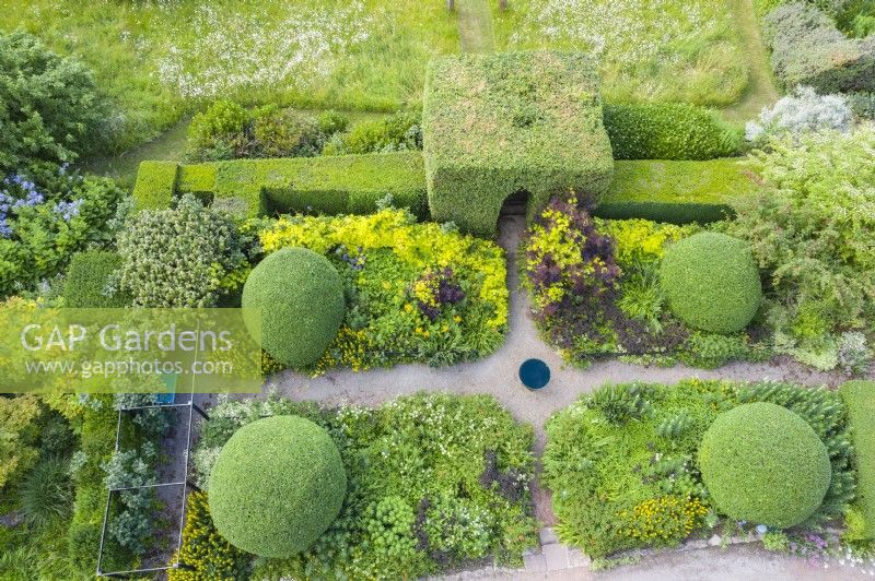 View over small formal garden backed by hedge of Box and hornbeam tunnel. Herbaceous plants with gravel paths, large clipped mounds of Osmanthus burkwoodii and  a bird bath. Image taken with drone. June. Summer. 