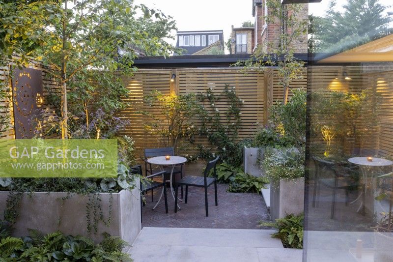 Courtyard garden at dusk with seating area, raised beds and contemporary wood boundary fence and lighting