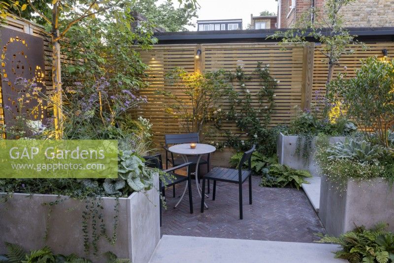 Courtyard garden at dusk with seating area, stone raised beds and contemporary wood boundary fence and lighting