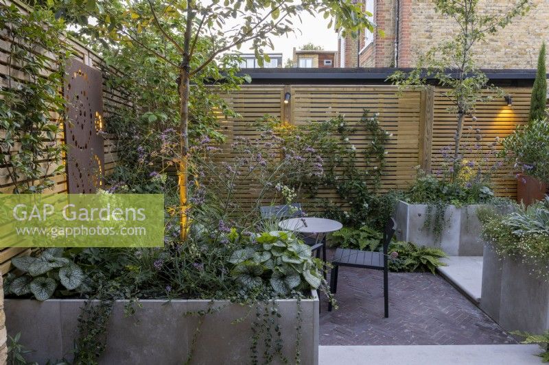 Courtyard garden at dusk with seating area with raised beds and contemporary wood boundary fence and lighting
