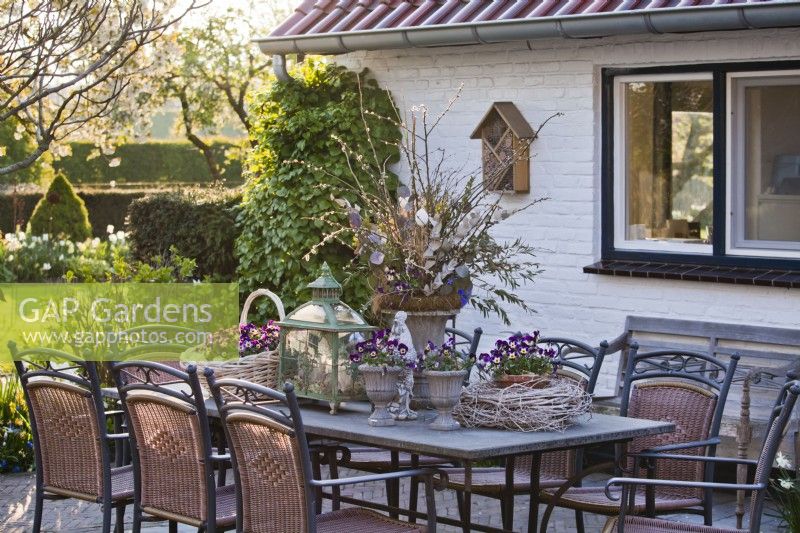 Outdoor table arrangement featuring decorative urns, basket and pot planted with Violas, lantern and wreath of twigs.
