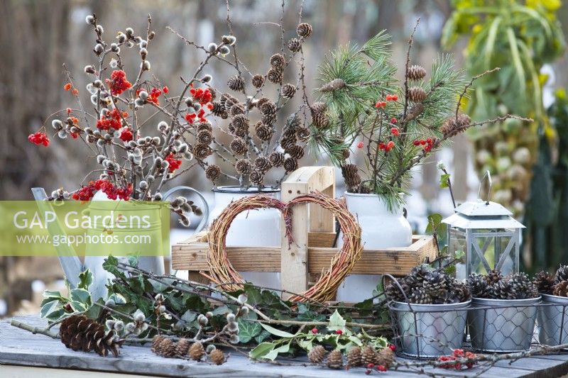 Winter arrangement featuring bunch of pussy willow and guelder rose berries in a watering can, wrapped heart wreath and pine and larch twigs with cones in milk churns.