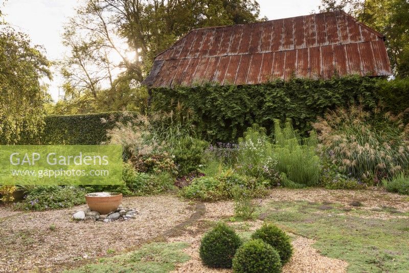 Gravel garden with bubbling water feature in front of barn with corrugated metal roof, at Terstan in September